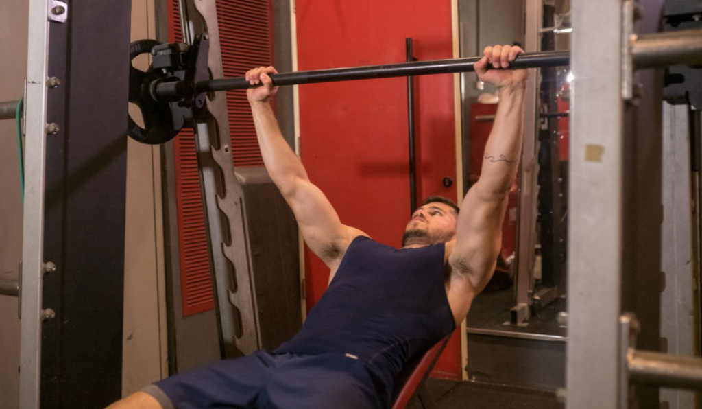 גבר שוכב על ספת חדר כושר מחזיק מוט במכונת הסמית' עם ידיים ישרות