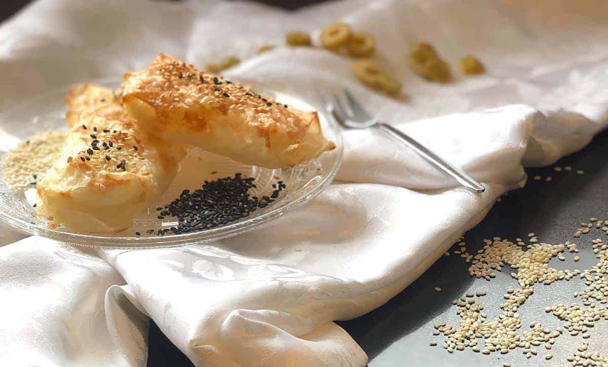 בורקס עשוי מדפי אורז על מפה לבנה ולידו חתיכות של זיתים, סומסום מפוזר ומזלג