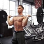 גבר צעיר וחטוב מחזיק מוט על הכתפיים מקדימה עם משקל בלי חולצה