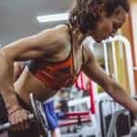 אישה צעירה בחדר כושר עם טופ מחזיקה משקולת ביד ימין ונשענת קדימה על מדרגה