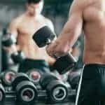 גבר שרירי עומד עם הגב למצלמה מחזיק משקולת יד מסתכל במראה