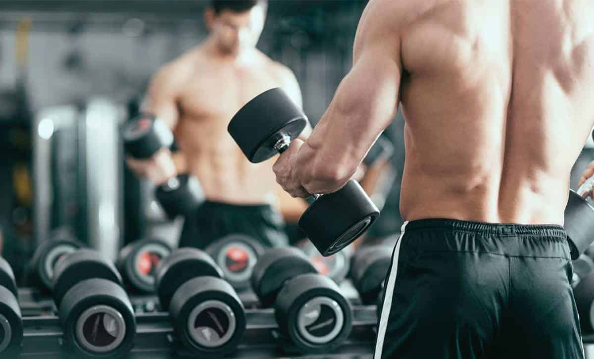 גבר שרירי עומד עם הגב למצלמה מחזיק משקולת יד מסתכל במראה